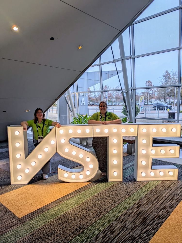 Two women stand behind giant lighted VSTE letters