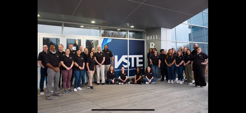 A group of about 25 people in matching black shirts pose for a group photo in front of the VSTE logo