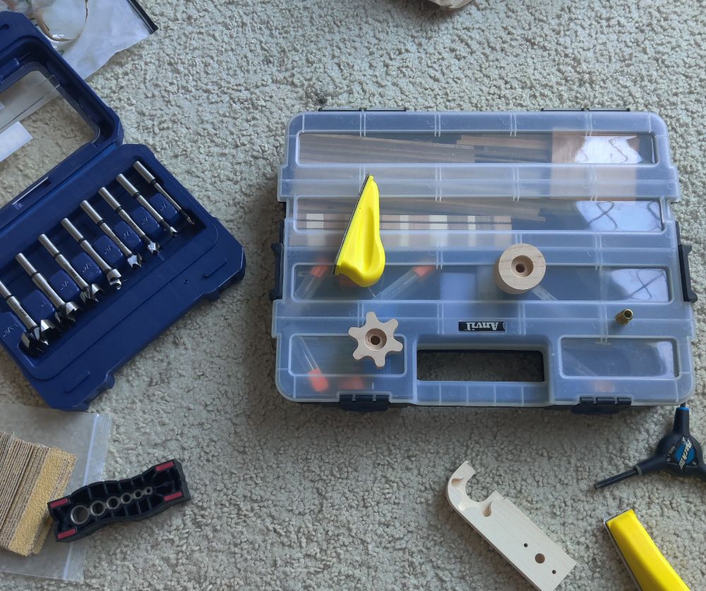 A collection of woodworking tools and parts spread out on a beige carpet. A blue case of Forstner drill bits is open on the left, and a transparent plastic storage box holds various CNC drill bits. On top of the storage box, there is a gear-shaped wooden piece, a round wooden piece, and two yellow plastic mini-sanders. Nearby are sandpaper sheets, a black plastic drill jig, a hex key tool, and a carved wooden handle with notches that match the gear-shaped piece.