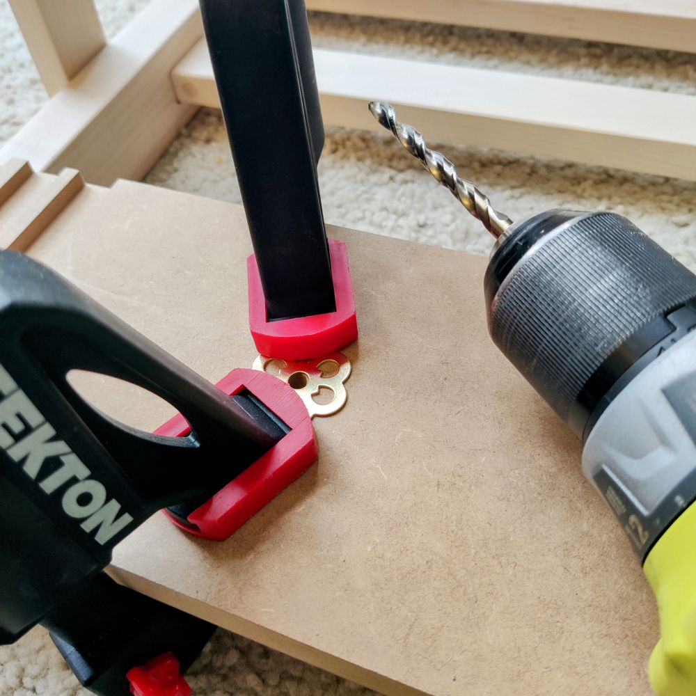 A woodworking setup shows a decorative flower-shaped brass washer secured to MDF by two bar clamps. A power drill with a spiral drill bit is positioned nearby, ready to bore a hole.