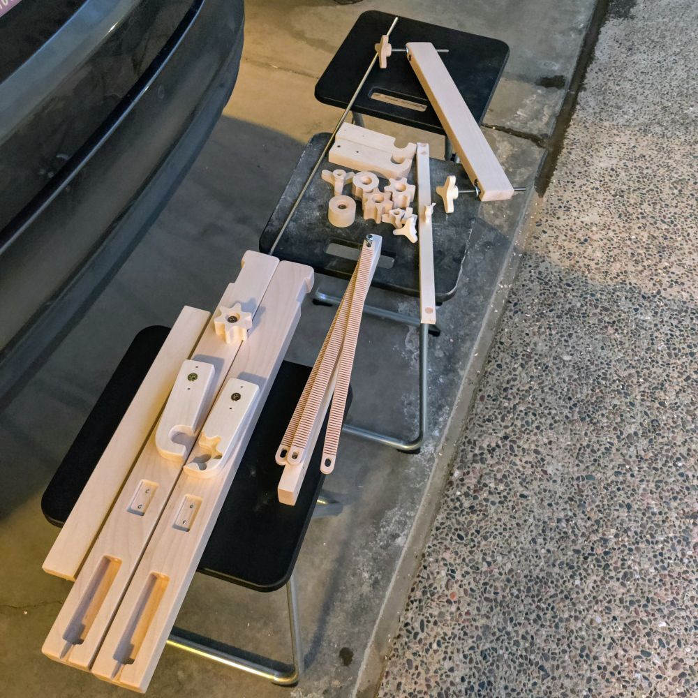 A collection of freshly sanded wooden parts laid out on two stools in a garage, including slotted beams, pegs, gears, and various shaped components for assembling a tapestry weaving loom.