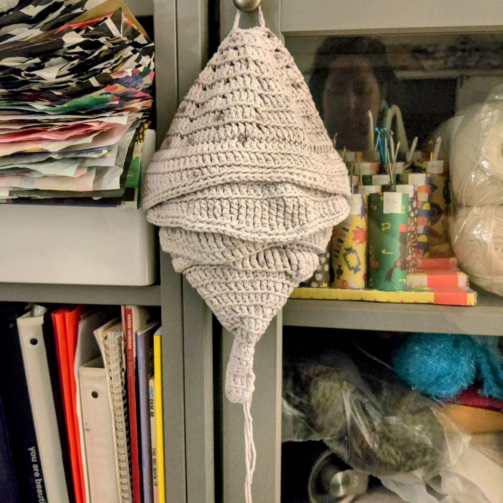 After bleaching and dyeing: The same crocheted hornet’s nest now appears in a speckled gray color, hanging from a knob on a gray cabinet. The textured yarn layers remain visible, and the background includes stacks of papers, craft supplies, and skeins of yarn.