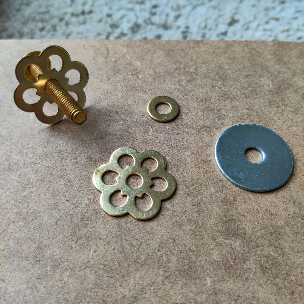 An assortment of washers laid out on a piece of MDF. There's a brass threaded bolt fitted with a decorative flower-shaped brass washer, a small brass washer, a large silver-colored zinc washer, and another brass flower washer beside them. The washers are arranged in a way that highlights their different sizes and shapes, with the decorative flower washer standing out due to its intricate design.