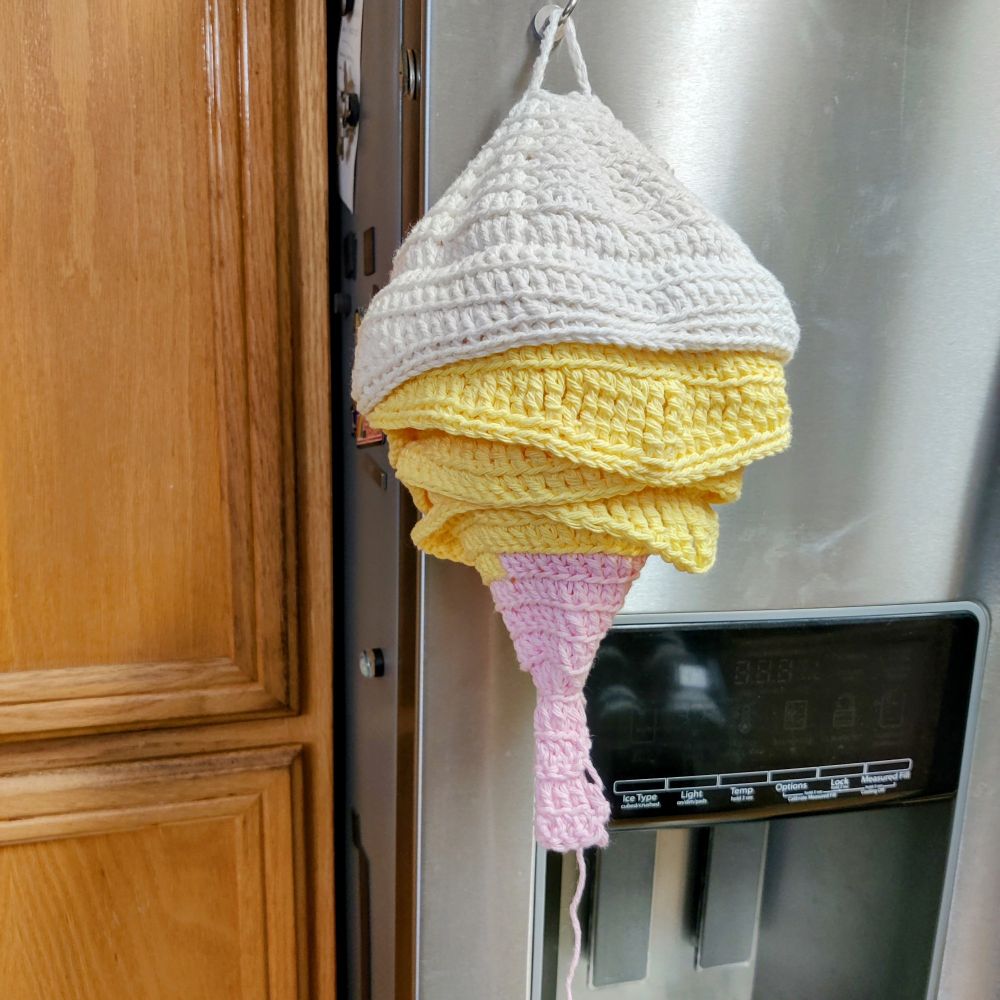 Before bleaching and dyeing: A crocheted imitation of a hornet’s nest, designed with pastel-colored yarn, hangs from a metal hook on a refrigerator. The layers resemble soft-serve ice cream, with pink at the bottom, yellow in the middle, and white on top.
