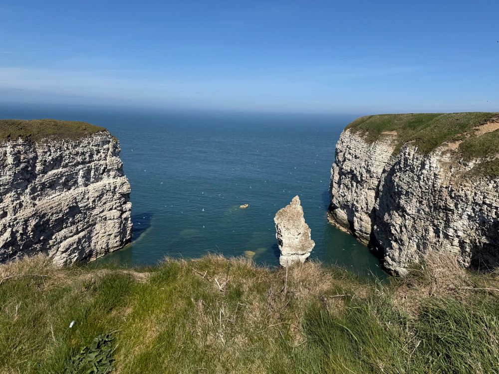 Blue sky, sea. Grass clifftop in the foreground. There are cliffs on both the left and right of the photo, as the camera is looking down into a small cove. There’s a big rock sticking up out the middle of the sea. The cliffs are white and topped by grass.