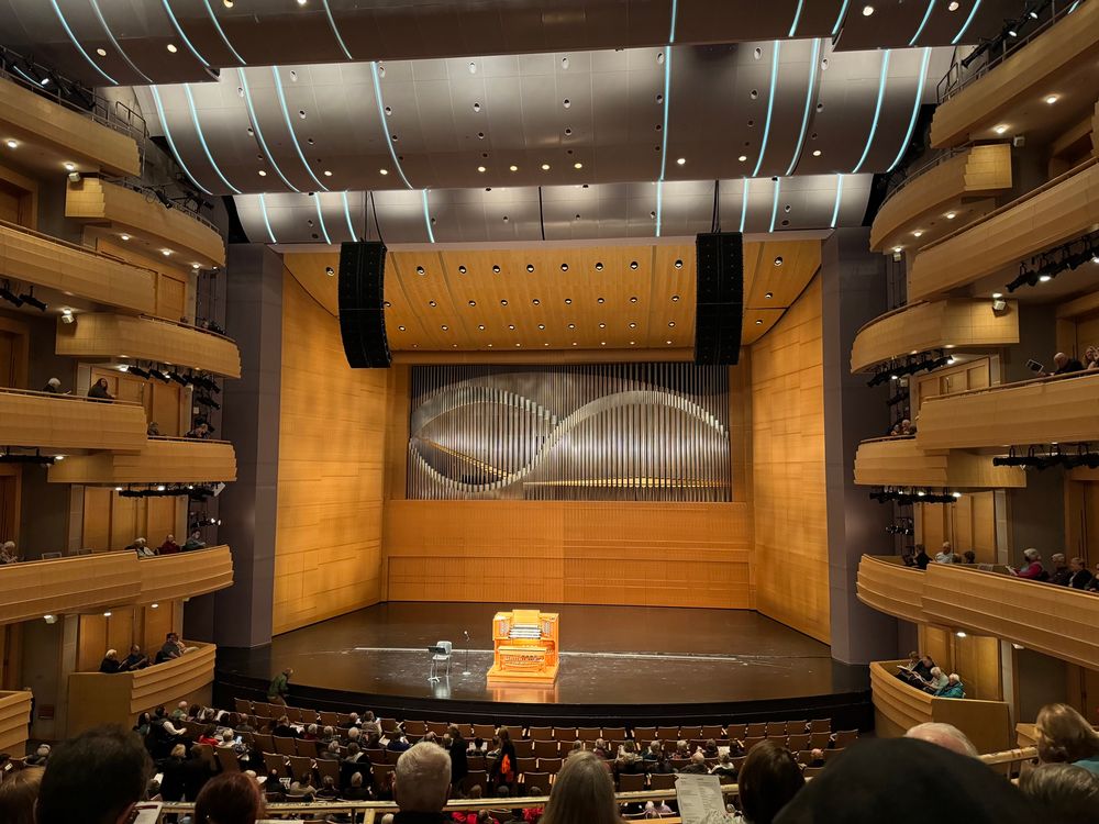 An organ console, a chair, a music stand and a microphone on an otherwise empty stage. Above and behind the stage are the organ pipes, arranged in two overlapping sine curves to great effect. There are modern balconies to either side. 