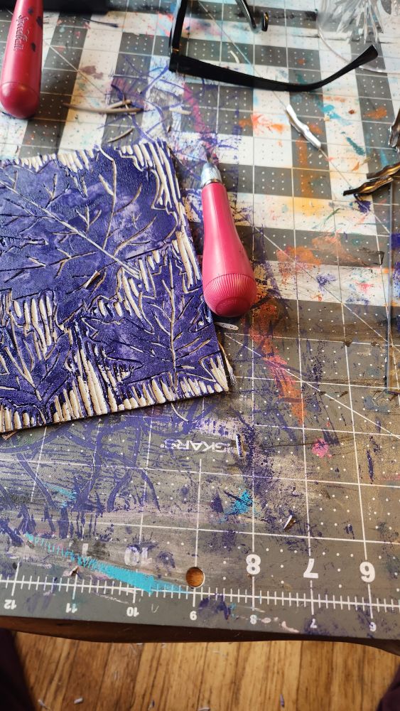 cutting mat with a linoleum carving tool and a pice of linoleum carved with leaves on it
