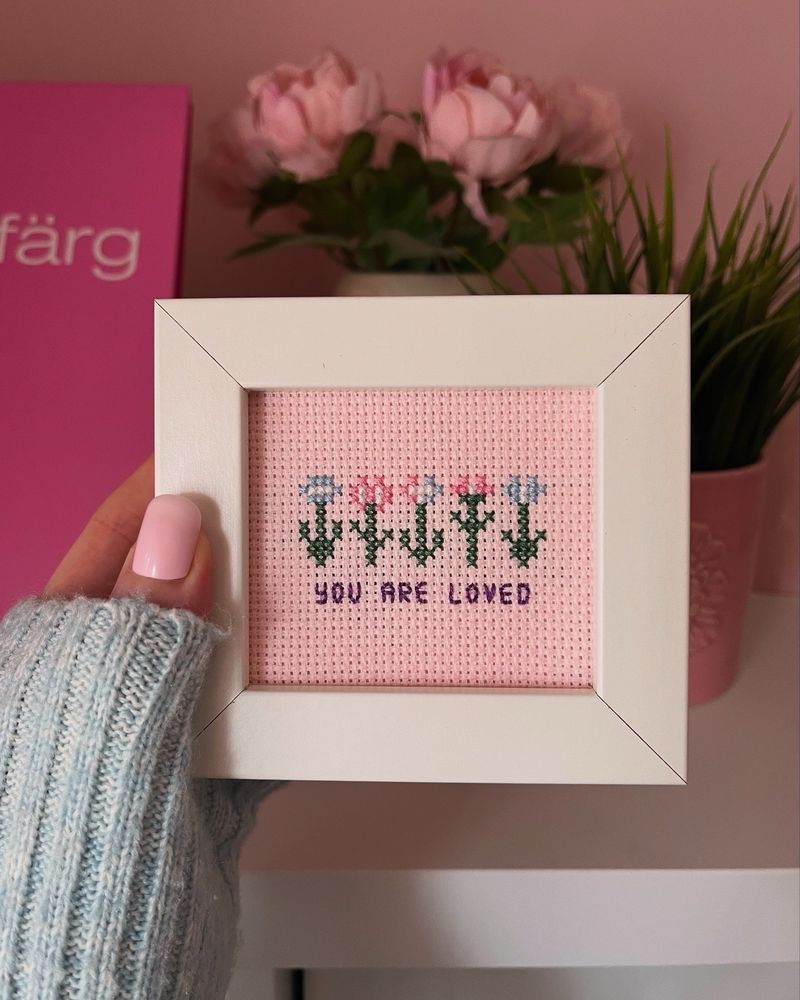 A picture of a small cross stitch artwork featuring pixel flowers in the colours of the trans flag, along with the text ‘you are loved’ in purple