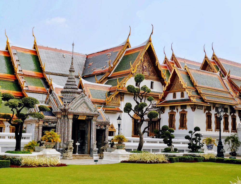 An ornate Thai palace with many steep roofs. The main part is white and the roof is green surrounded by orange. There are topiary trees in front and some bright green grass. 