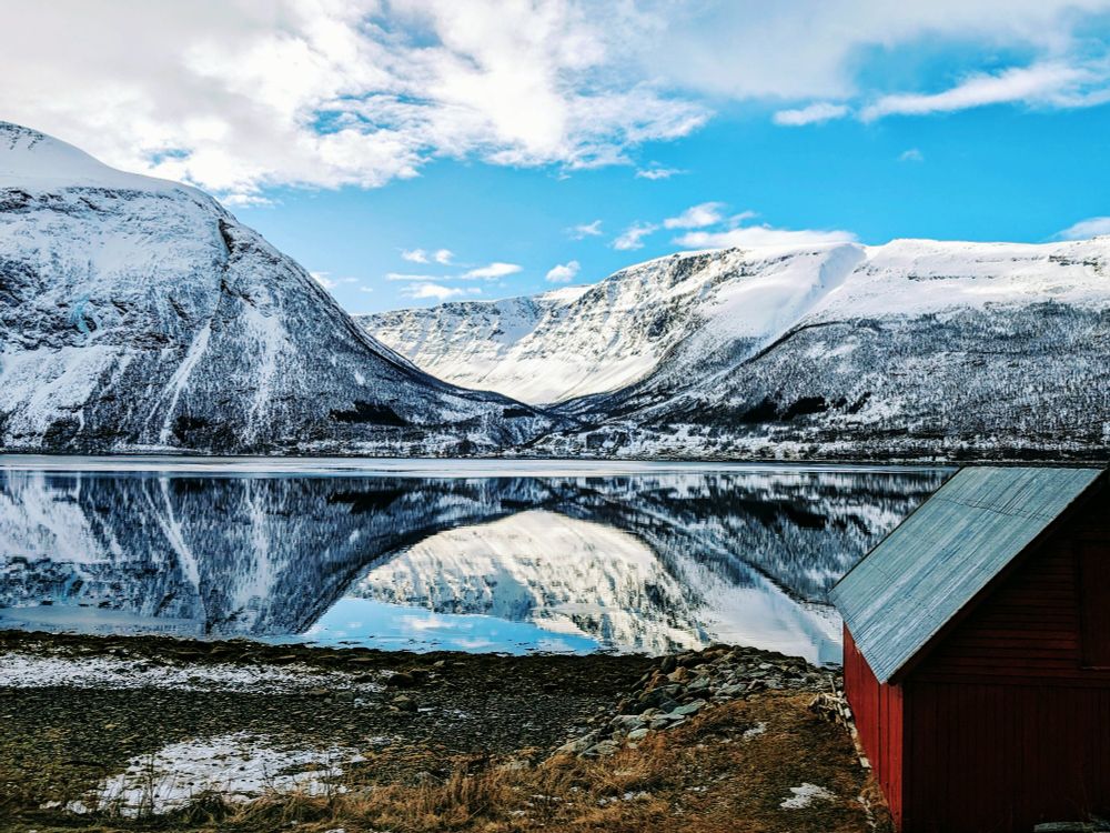 Snow-covered mountains reflected in perfectly smooth water with a bit of brown grass with patches of snow in the foreground. There's part of a little red cabin with a metal roof in the right corner. The sky is bright blue with thick clouds at the top. 