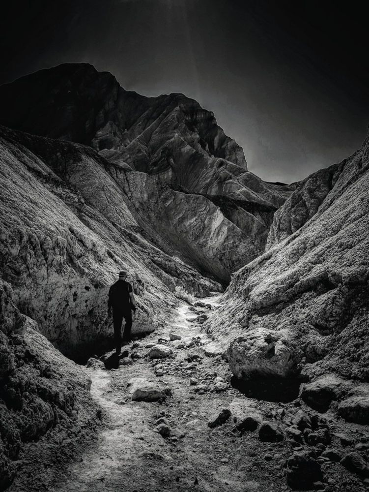 A black and white photo of a man walking through a barren canyon. The sky is nearly black. 