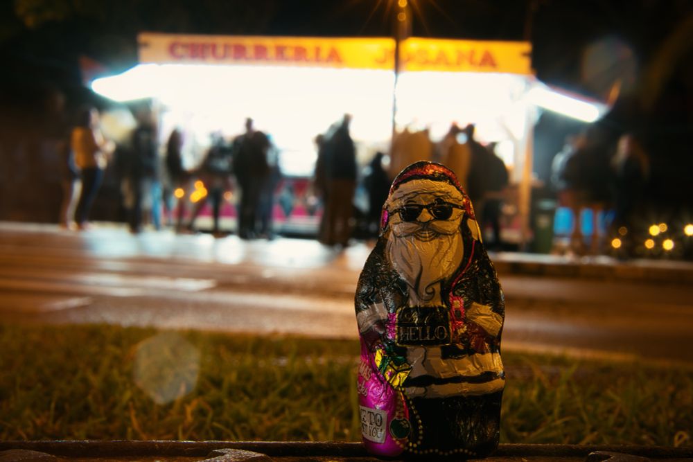 Ein Mini-Schoko-Weihnachtsmann steht bei Nacht vor einer hell erleuchteten Churreria, vor der viele Menschen anstehen 