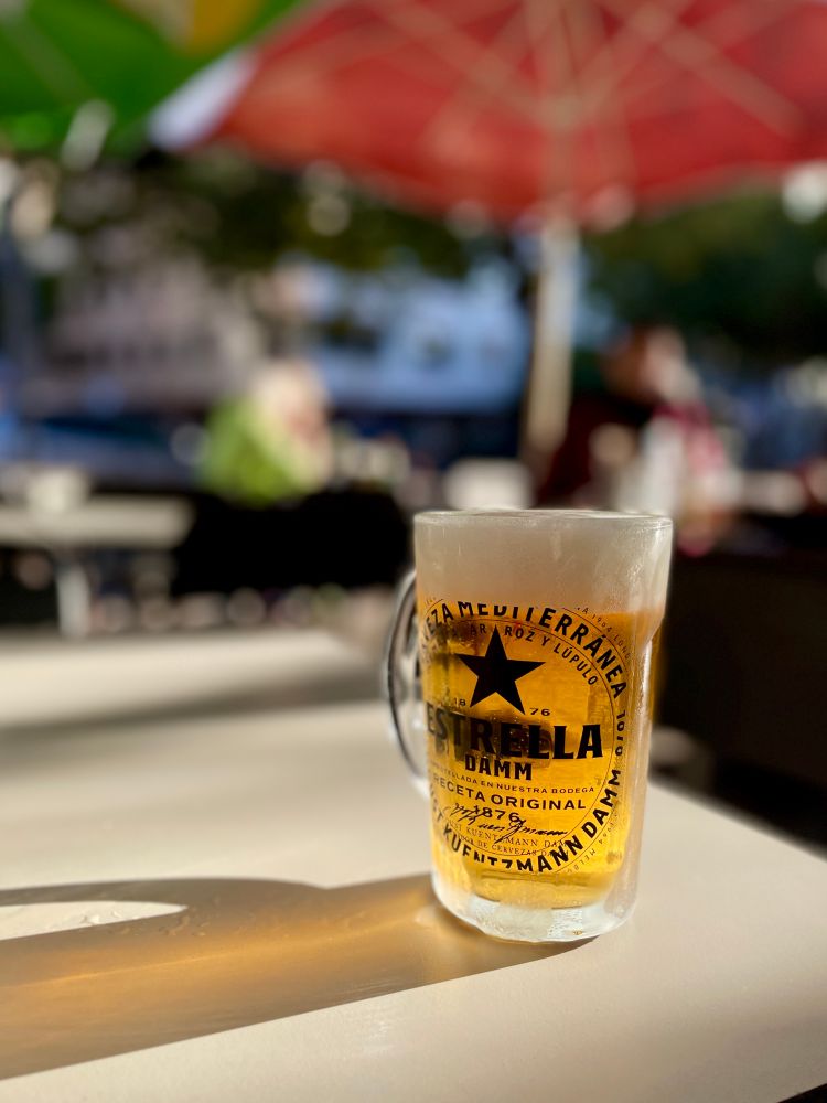 Ein von der Abendsonne angeschienener Glaskrug mit goldenem Bier und einer Schaumkrone bis an den Glasrand. Nach links zieht sich ein langer Schatten auf einem weißen Tisch. Im Hintergrund unscharf in der Sonne sitzende Gäste