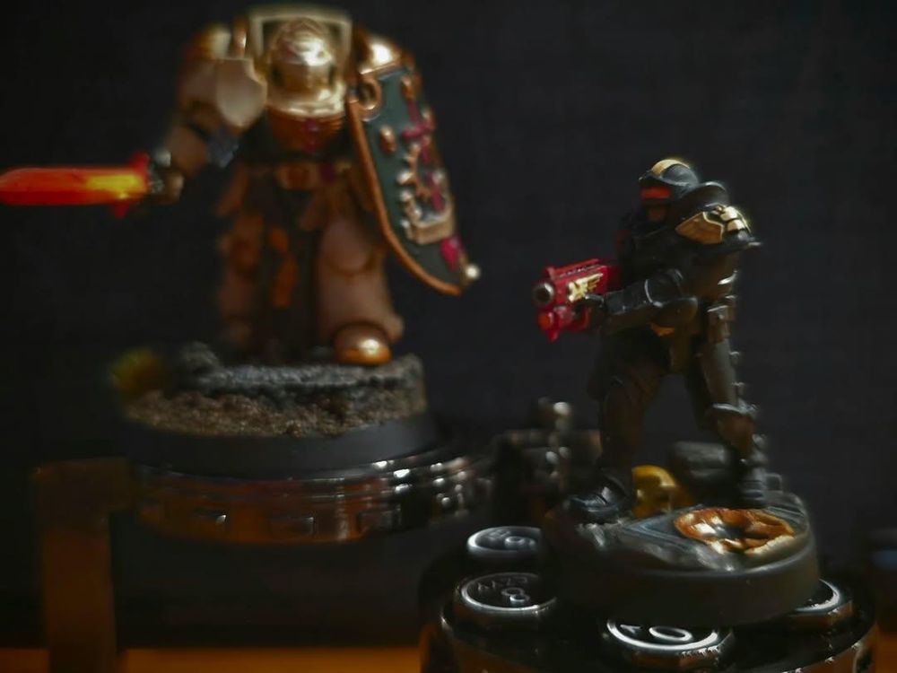 Two warhammer 40k models are pictured atop a novelty revolver dice set, an adeptus arbites model is in focus with an out of focus Deathwing Knight behind it 
