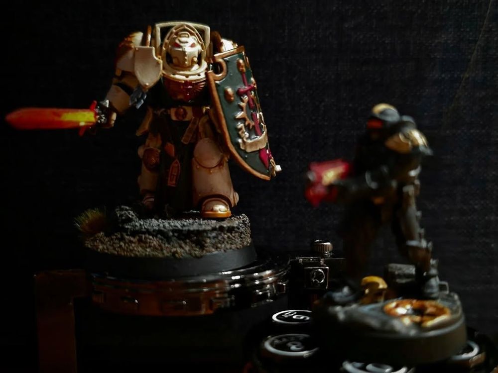 Two warhammer 40k models are pictured atop a novelty revolver dice set, an Deathwing Knight model is in focus with an out of focus Adeptus Arbites model in the foreground  