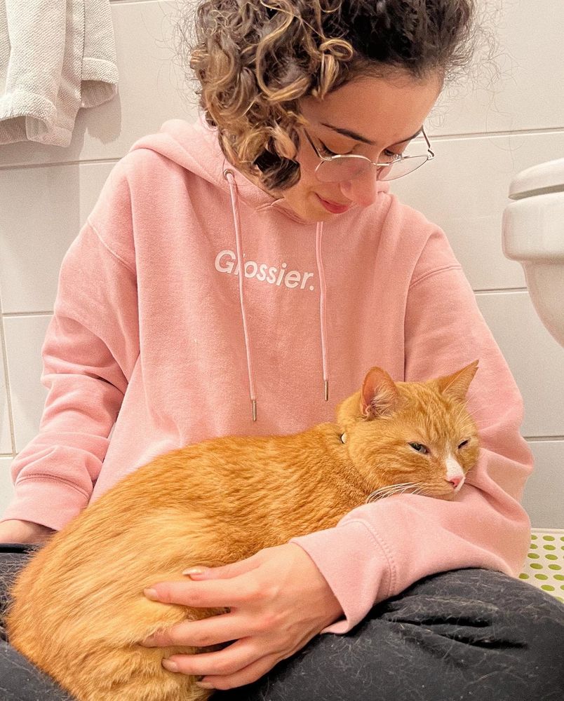 me wearing a pink glossier hoodie, looking down at my orange cat smartie in my lap