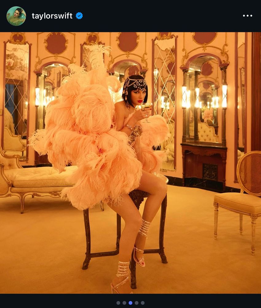 Instagram post by Taylor Swift. She's wearing a dark wig, a bob with bangs, and a big feathery showgirl outfit in a fancy dressing room holding a drink.
