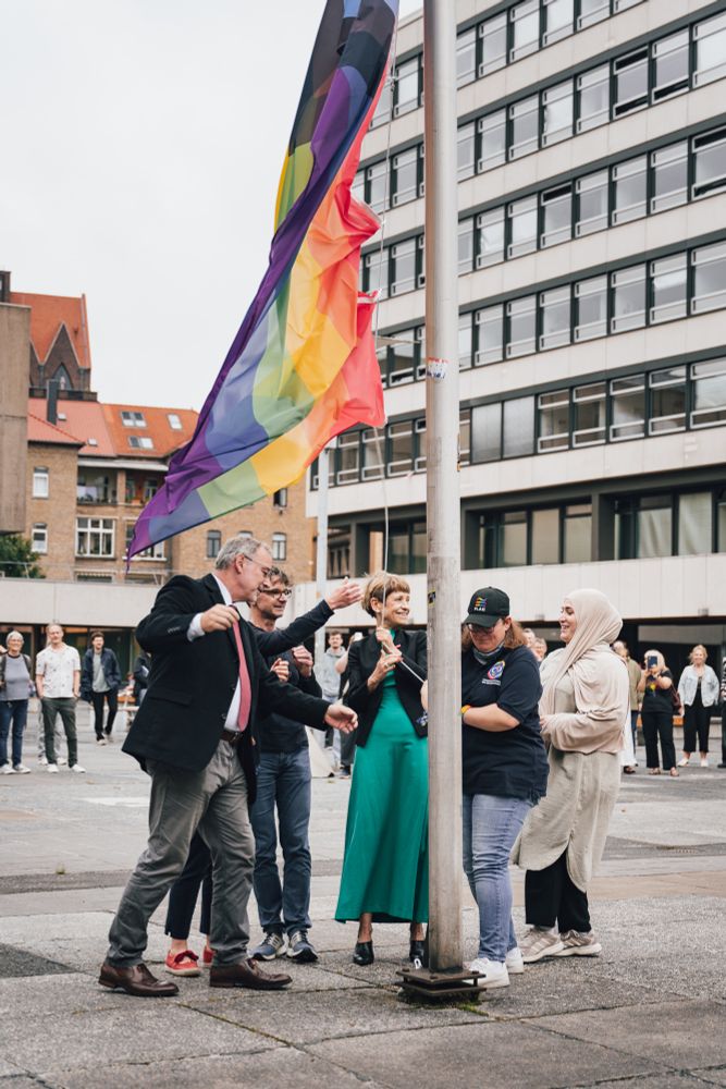 Regenbogenflagge weht am Forumsplatz der TU Braunschweig. Menschen stehen gemeinsam, halten inne. Im Hintergrund Gebäude der Universität.