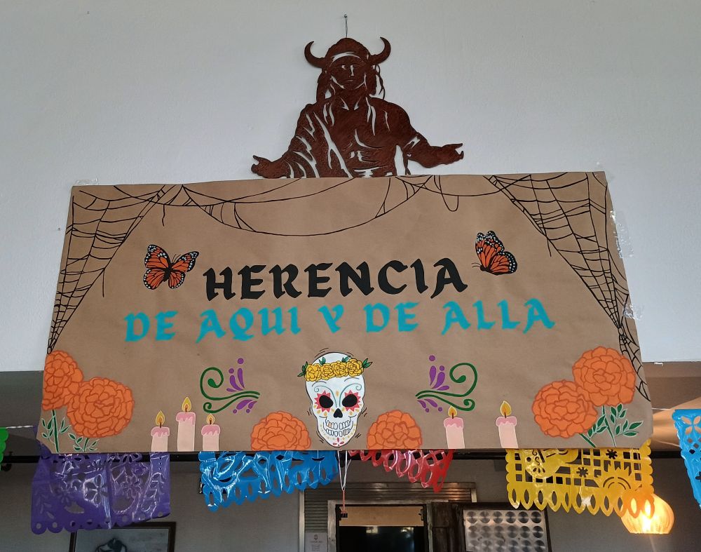 A sign that says Herencia, de aquí y de allá. With Dia de muertos decorations 
