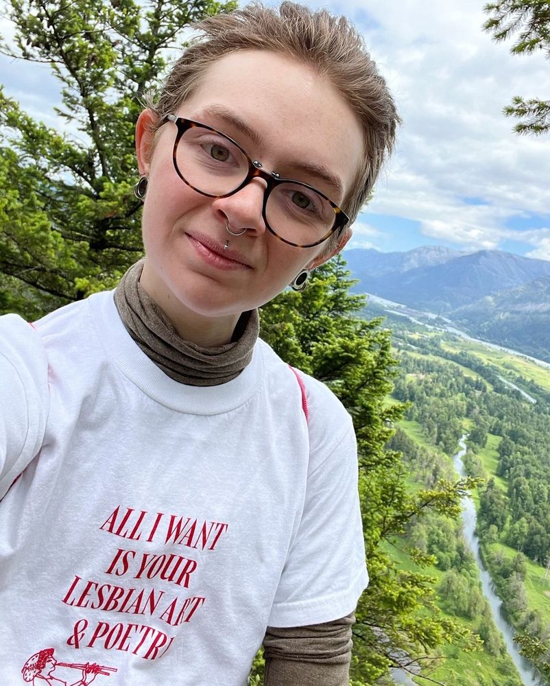A picture of Cavar, a genderless person with short blonde/brown hair, glasses, and visible facial piercings, standing on a mountain with a picturesque view behind them.