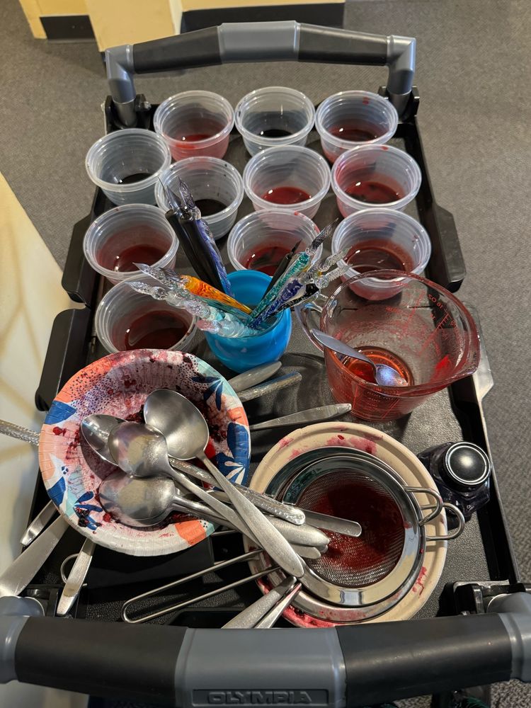 A messy cart full of small cups of red and black liquid, glass dip pens, spoons, and bowls filled with berry juice and pulp.