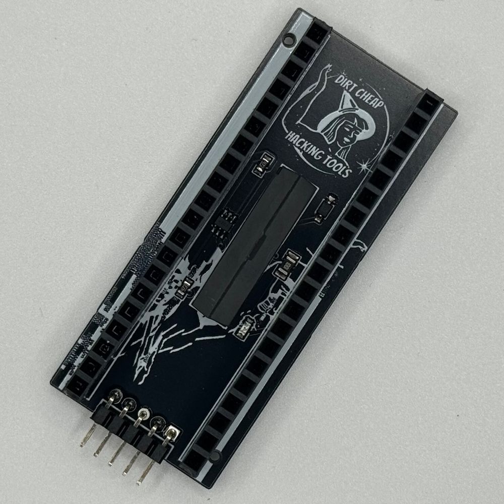 black and white circuit board with a dirt cheap hacking tools logo and glitchy solder mask art and 7 components, 2 large rows of female dupont headers, and perpendicular a row of 5 90 degree dupont male headers 