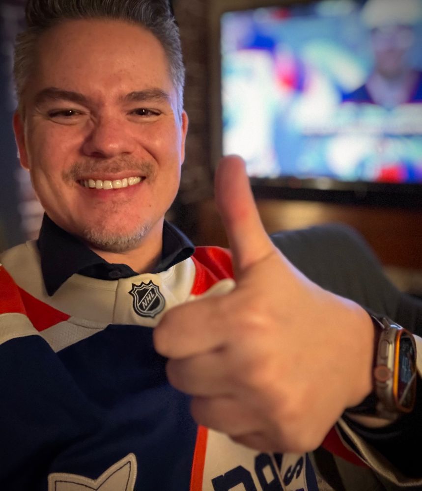 Paquette in an oilers jersey giving the thumbs up with the game in the tv behind him.