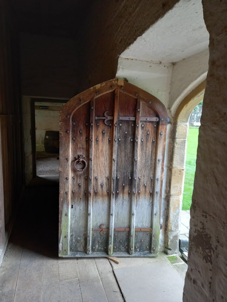 Sturdy wooden door with iron ring handle and rivets. Dimly lit corridor leading to a covered walkway in background.