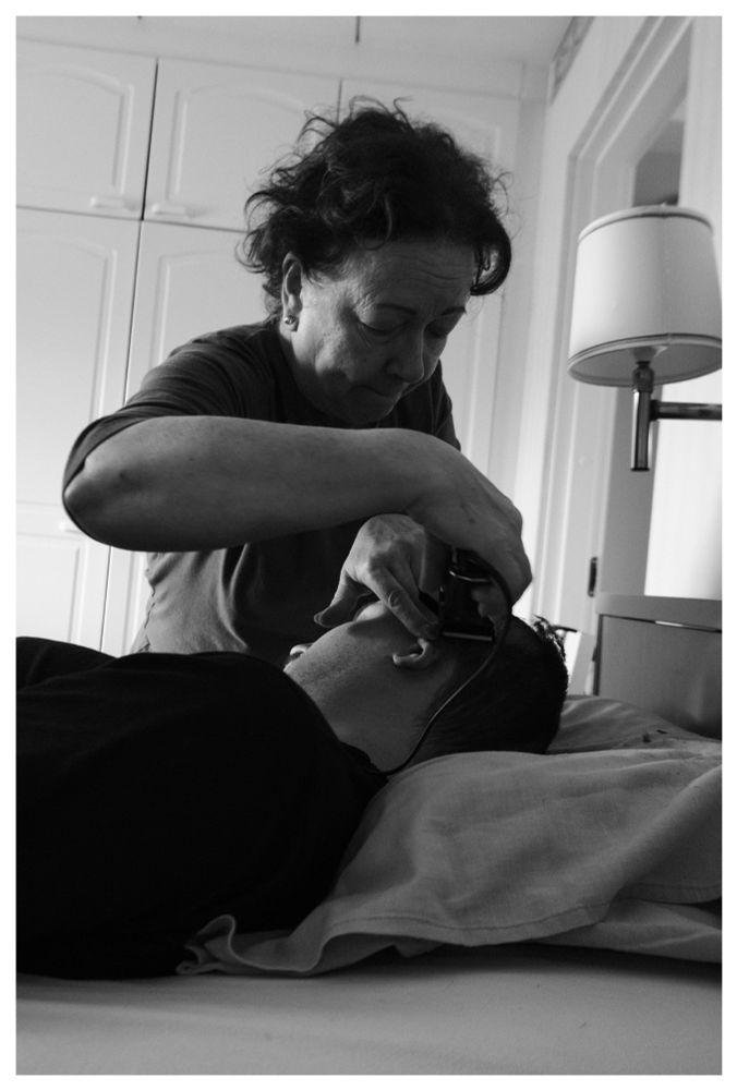 A black and white image. The author of the post is laying in bed, you can see his head and shoulders. He is in the lower half of the photo. There’s a pillow under his head. His face is turned away from the camera. Behind his back, his mother is using hair clippers to cut his hair short. They are cutting the side of his head. The mother is leaning over him. 