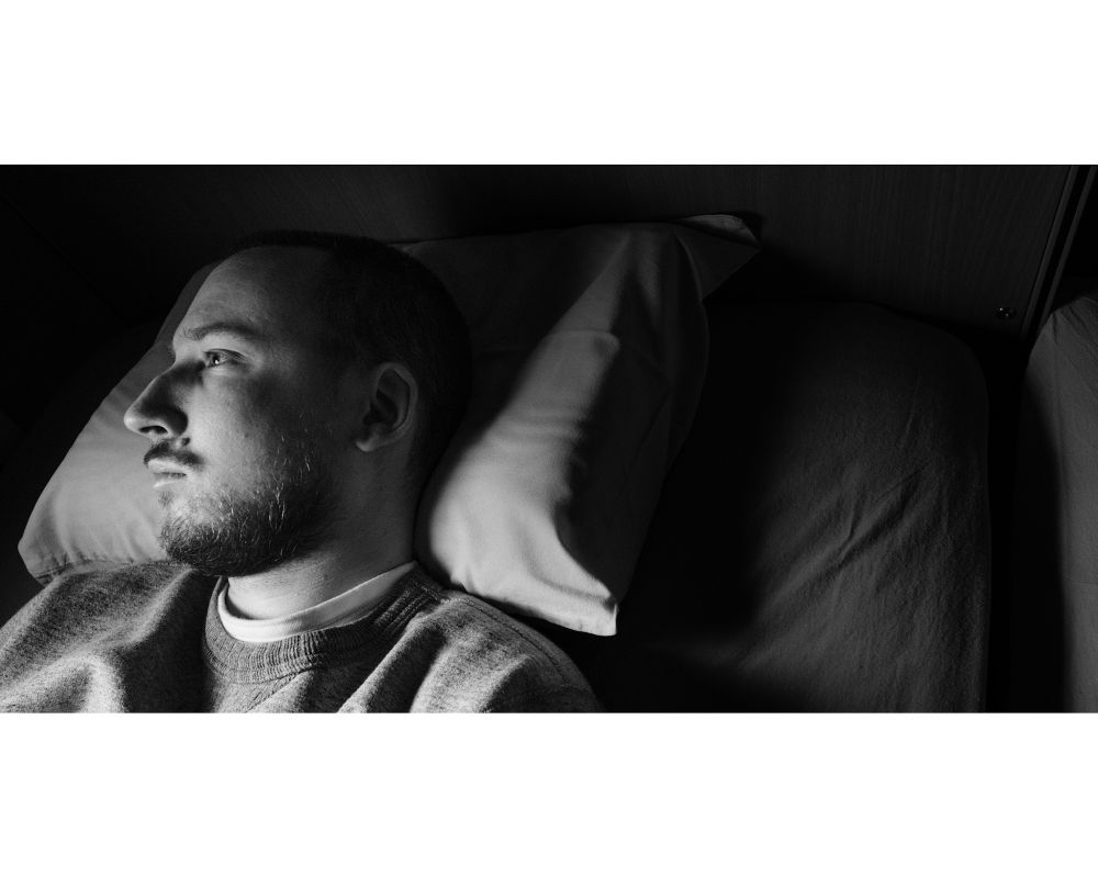 A black and white image. The author of the post is laying in bed, he’s a man in his 30s. He has a beard and a moustache. You can see his head and part of the shoulders. His head is turned sideways and his looking at the distance. The picture is dark, the harsh light is hitting his head and lighting his facial features, like his Roman nose. He is positioned in the left side of the photo and is looking at that direction. Behind his head is a lot of negative space, there is a part of his pillow, that is under is head, and part of the bed is shown. 