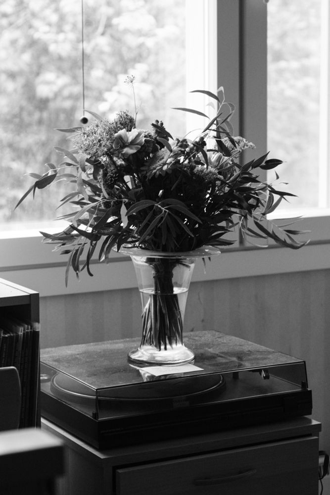 A black and white photo of a flower bouquet on a record player, next to a window that illuminates natural light inside the room.