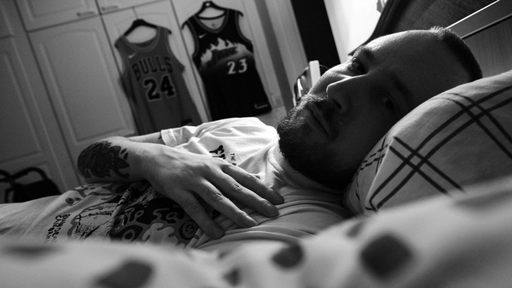 A black and white photo of Niko, a man with a dark beard, lying in bed. In the foreground a pillow is shown. Middle ground is Niko with his head on a pillow, looking at the camera, having his hand on his cheast. Background shows white cabinet doors and two basketball jerseys hanging on them.
