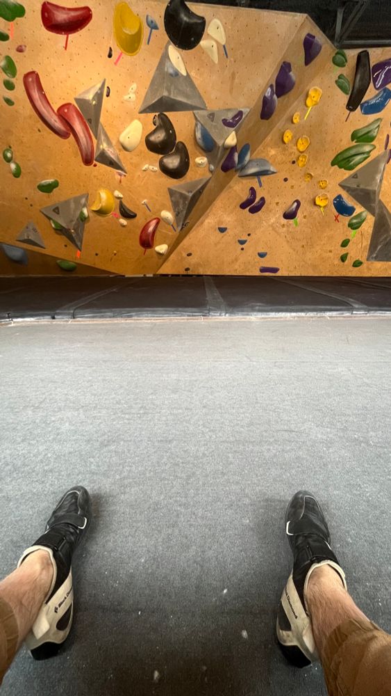 A point of view of a man sitting in a bouldering gym. His feet are outstretched in front of him, with black and white climbing shoes on his feet. The gym floor and mats stretch out in front, and in the background is the climbing wall with holds and problems of differing difficulty.