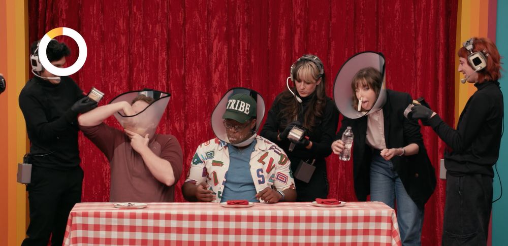 Three members of Dropout's Game Changer production design team funneling Parmesan into players' mouths/neck cones.