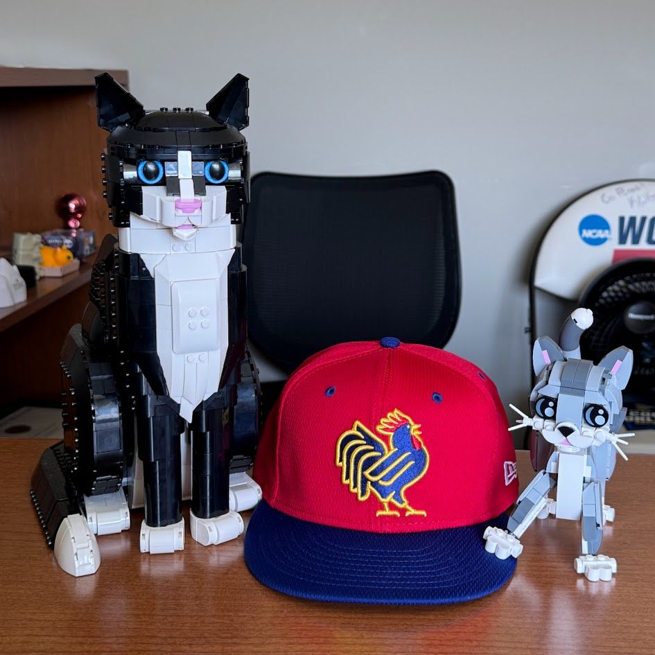 A red and blue Northwest Arkansas Naturals batting practice cap on my desk with HATS the Lego Cat and CAPS the Lego Kitten
