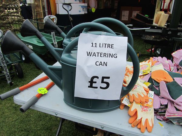 Green watering can displayed next to a pile of gloves on a stall selling gardenalia. There's a sign attached to it saying "11 litre watering can £5".