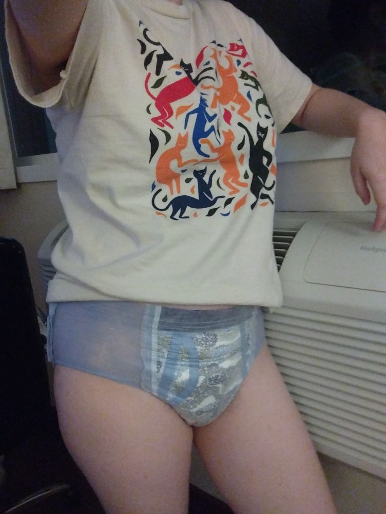 someone taking a photo while standing in front of a window. their arm is resting on the windowsill, and they are wearing a blue goodnite. their shirt has many colourful dancing cats arranged in a square.