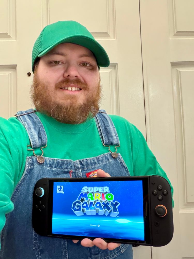 Me dressed as Luigi, with a green long sleeve t shirt, denim overalls, and a green ball cap. I’m holding a Switch 2 with the Super Mario Galaxy title screen displayed.