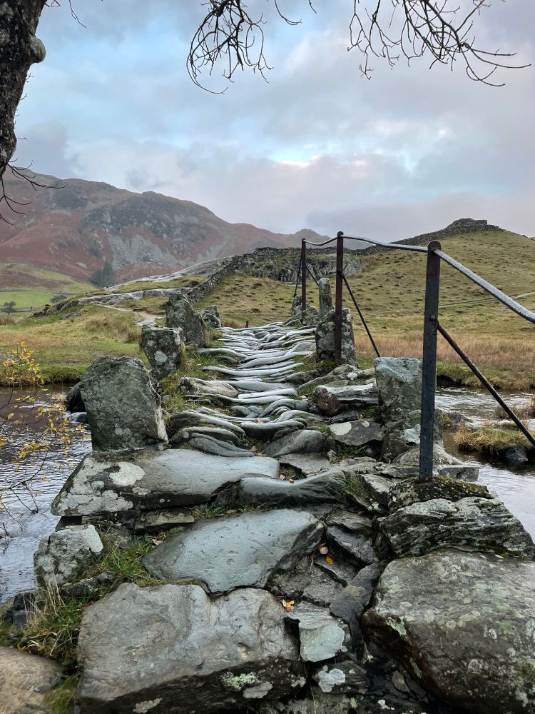 A cobbled bridge over a small river with mountainous terrain in the background