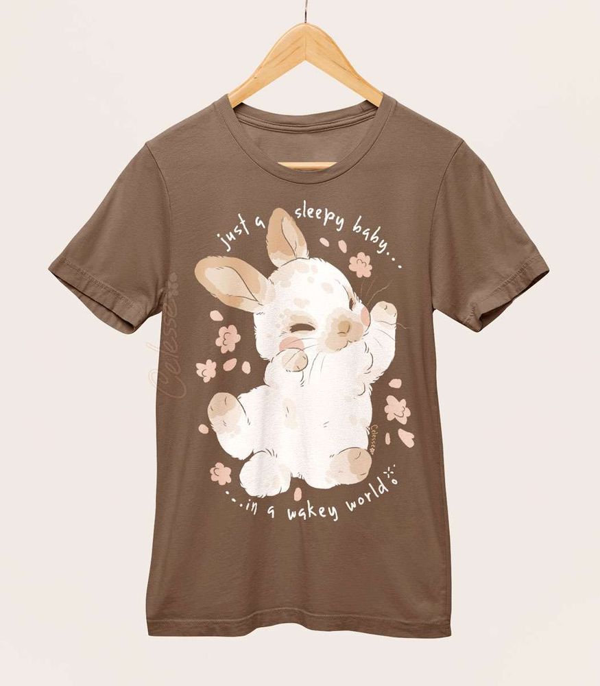 A brown shirt on a wood hanger with a print featuring a sleepy baby bunny sprawled out and the text "just a sleepy baby...in a wakey world."