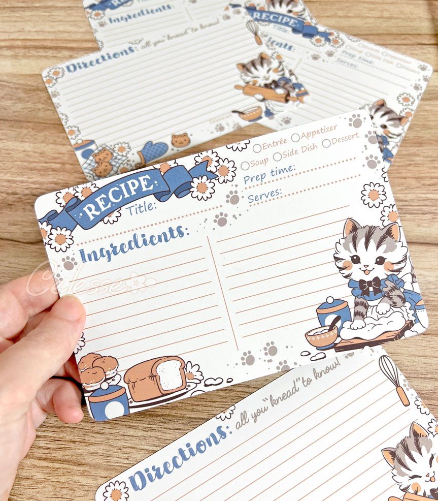 A photo of a hand holding up a recipe card over a wooden tabletop, several more copies of the card scattered in the background. The print features vintage style art of a kitten in a frilly dress and apron kneading dough, several baked goods and white daisies framing the note lines of the card. The back side features a section for directions with the line "All you 'knead' to know!"