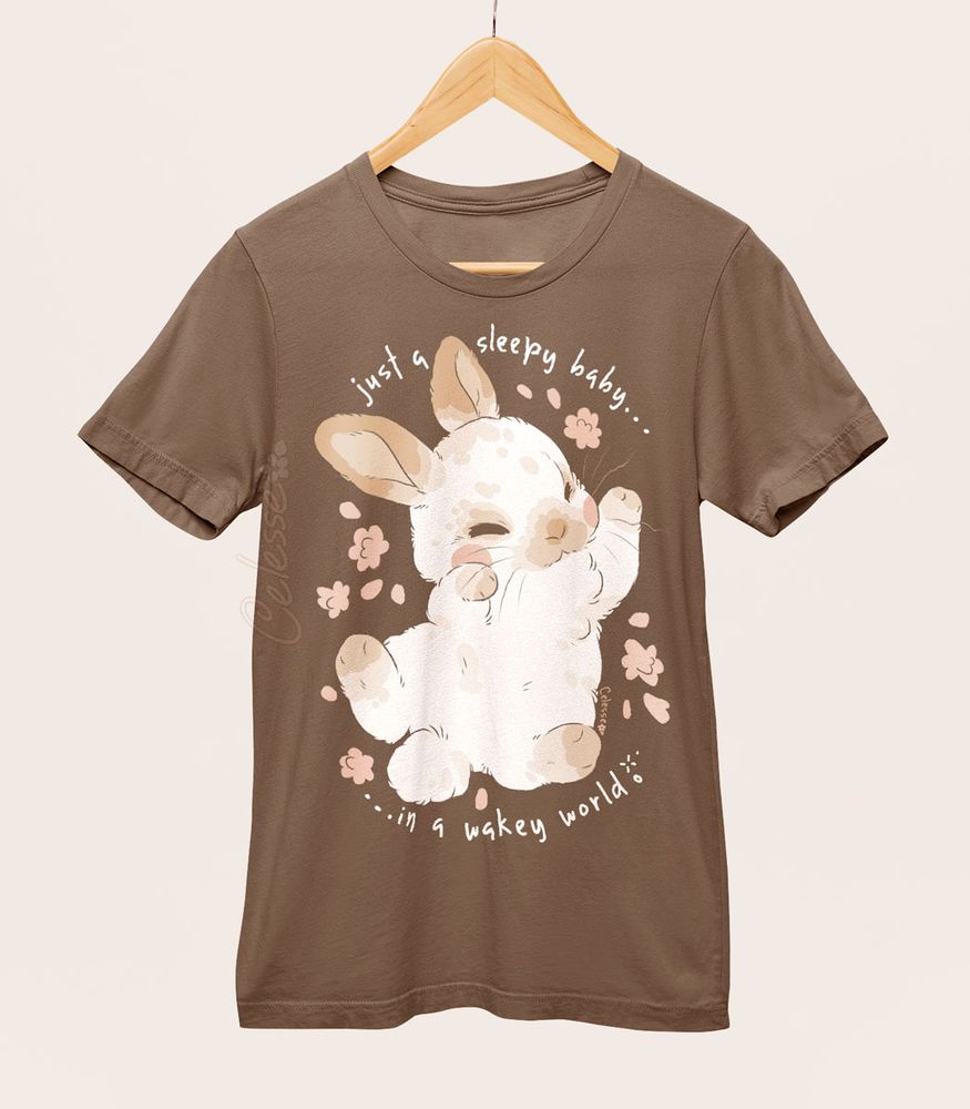 A photo of a t-shirt hanging from a wood hanger. The shirt is a soft muted brown and the print features a sleepy baby bunny sprawled out with flowers around it. The text reads "just a sleepy baby...in a wakey world."