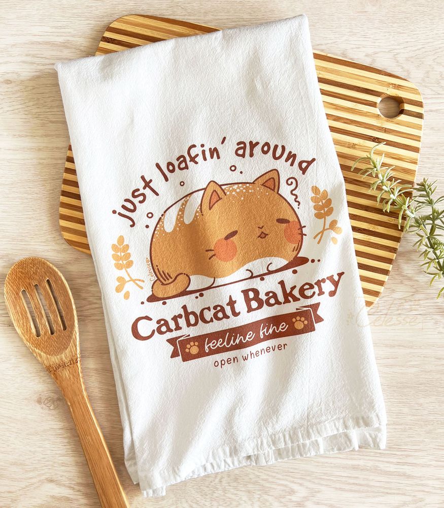 A photo of a white tea towel featuring a bread loaf cat logo called Carbcat Bakery, laying on a wood table on top of a lined cutting board, a wooden spoon laid next to it. The extra text reads "just loafin' around" and "feeline fine".