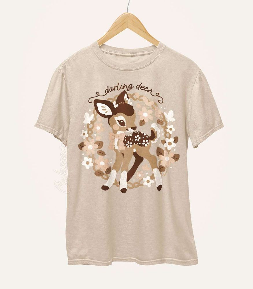 A tan shirt on a wood hanger with a print featuring a baby deer in a wreath of flowers with the text "darling deer."