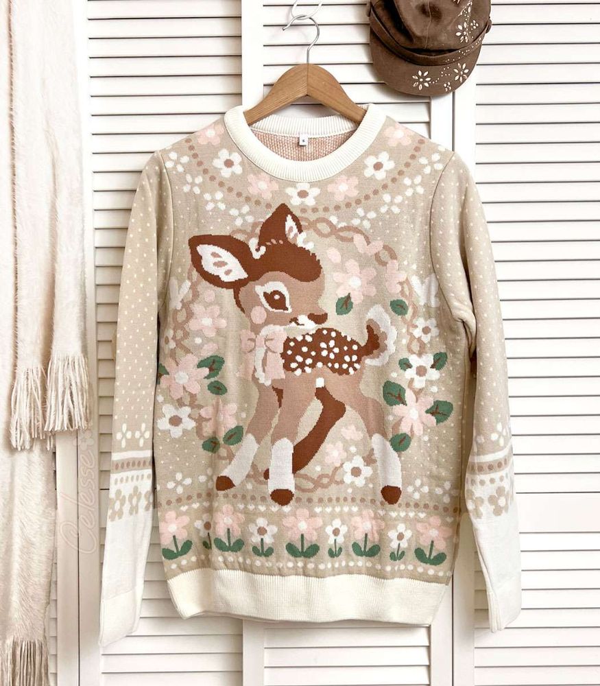 A photo of a knit sweater hung from a wood hanger on a white slat wall door. The colors are soft beige and browns and the front design features an image of a baby deer standing in a floral wreath.