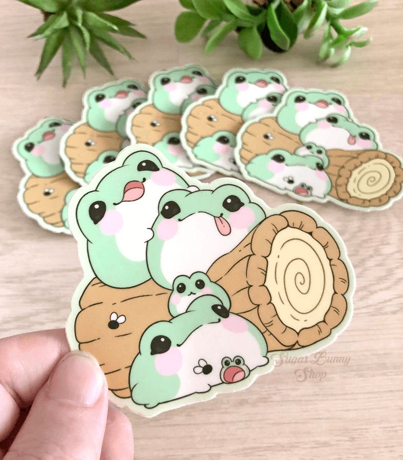 A photo of a hand holding a vinyl sticker that features round, fat blob frogs sitting on a log. On the wood table in the background are more of the stickers fanned out on display.