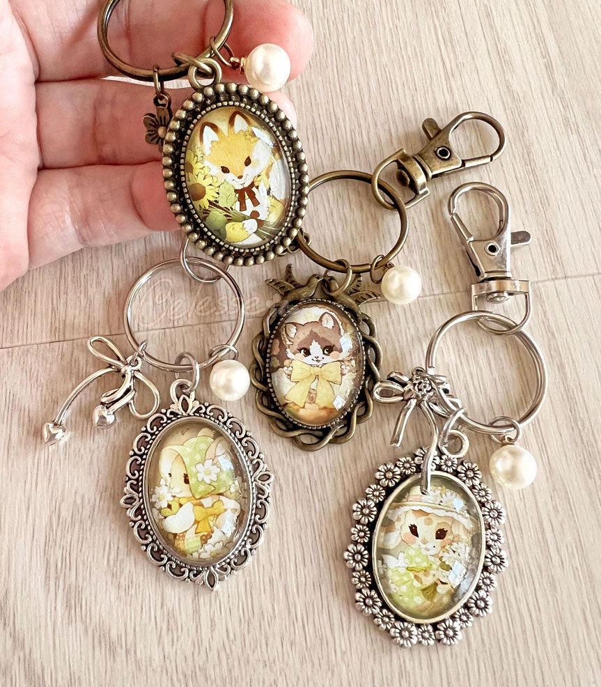 A photo of several vintage-style cameo keychains with intricate frames and dangling charms laid out on a table, a hand in the corner holding one up. The images inside the cameos are drawn portraits of various animals in old-fashioned clothing.