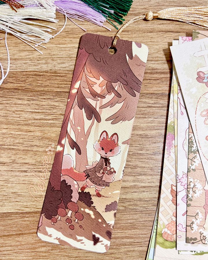 A photo of a paper bookmark laid on a wooden tabletop, the art featuring a fox wearing a simple old-fashioned dress and walking through a sunlight forest. The bookmark is made of thick textured paper and has rounded corners and a tassel attached at the top. Some extra tassels and a stack of other bookmark designs are partially visible on the outskirts.