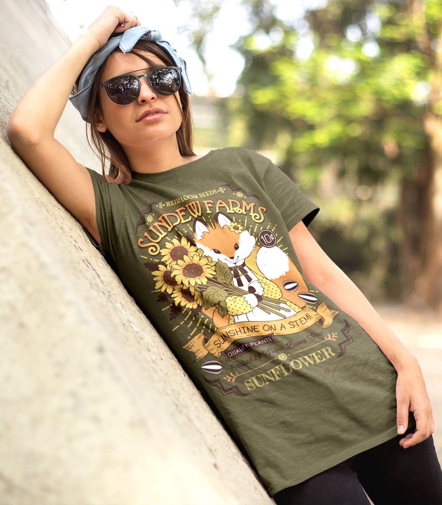 An angled photo of a person outside wearing sunglasses and leaning against a concrete wall. She wears an olive green shirt with a print of a vintage-style logo for Sundew Farms Sunflower Seeds, featuring a red fox wearing a yellow polka dot dress holding a bundle of sunflowers in their arms.