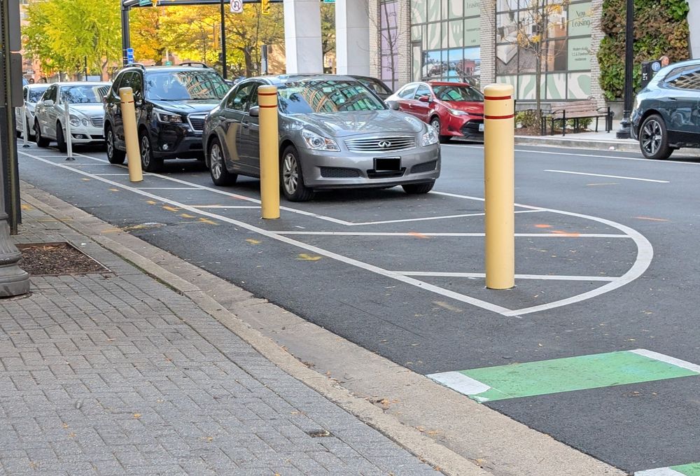 A row of three yellow bollards in the buffer area of a parking-protected bike lane. Traditional white flex posts can be seen further down the street. The location is Clarendon Blvd near N Courthouse Rd across from the Wendy in the Courthouse neighborhood.